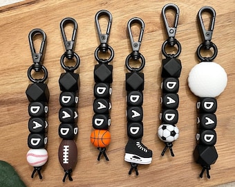 Könnte beinhalten: Sechs schwarze Silikon-Schlüsselanhänger mit dem Wort "DAD" auf jedem Anhänger. Jeder Anhänger hat einen anderen Sport-Anhänger: einen Baseball, einen American Football, einen Basketball, einen Fußball, einen Schlittschuh und einen weißen Kreis.