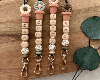 Puede incluir: Cuatro llaveros de cuentas de madera con detalles florales y etiquetas de nombre personalizadas. Cada llavero presenta un pequeño amuleto de maceta con una flor en la parte superior. Las etiquetas de nombre deletrean "MRS J", "MELL", "MISS K" y "MSG".