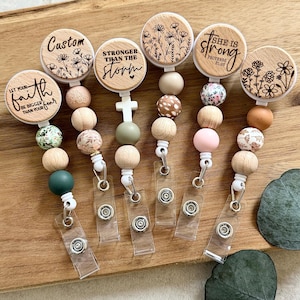 Könnte beinhalten: Eine Sammlung von Holz-Ausweisrollen mit verschiedenen inspirierenden Zitaten und floralen Designs. Jede Rolle verfügt über eine einzigartige Kombination aus Holz- und Dekorperlen sowie einen transparenten Kunststoffriemen mit Schnappverschluss.