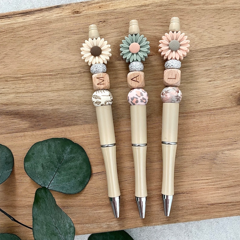 May include: Three beige pens with wooden beads and flower charms. The beads spell out the word "MAL".