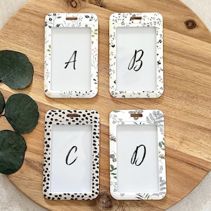 Puede incluir: Cuatro porta-credenciales blancos con estampados florales y d&aacute;lmatas. Cada soporte tiene una ventana transparente y una letra: A, B, C y D. Los soportes est&aacute;n sobre una superficie de madera.