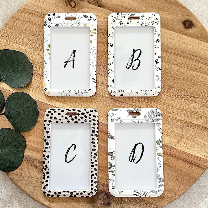 Puede incluir: Cuatro porta-credenciales blancos con estampados florales y d&aacute;lmatas. Cada soporte tiene una ventana transparente y una letra: A, B, C y D. Los soportes est&aacute;n sobre una superficie de madera.