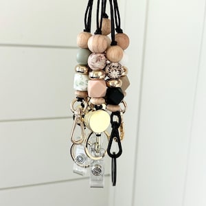 May include: A beaded lanyard featuring wooden, patterned, and gold-toned beads. It includes a retractable badge reel and a swivel clasp. The colour scheme incorporates natural wood tones, pink, black, and gold.