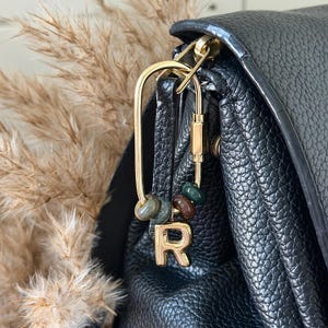 May include: A gold-toned metal carabiner clip attached to a black textured leather bag. The clip features three small, smooth, multi-coloured stone beads in shades of green and brown, along with a gold-toned metal letter R charm hanging from the bottom.