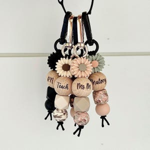 May include: Keyrings with black clips, wooden beads, and flower accents. The keyrings feature wooden beads with the words "Teach" and "Ms. M" printed on them. Other beads are in shades of pink, beige, and black.
