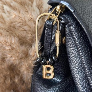 May include: A gold-toned metal carabiner clip attached to a black textured bag. The carabiner features a threaded locking mechanism and is adorned with four small, smooth black beads and a single gold-toned letter B charm hanging at the bottom.