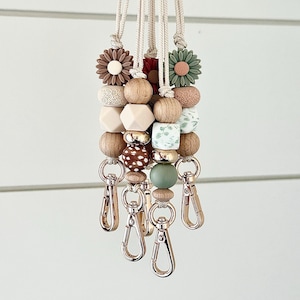 May include: Beaded lanyards in neutral tones, featuring wooden and silicone beads. Each lanyard has a unique design, including flower-shaped beads and gold-coloured metal clasps. The lanyards are suspended from a cream-coloured cord, suitable for keys or ID badges.