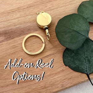 May include: A gold-coloured circular retractable badge reel with a matching gold-coloured split key ring. The text Add-on Reel Options! is printed in white script. The metal components have a smooth, reflective finish and a small black cord.