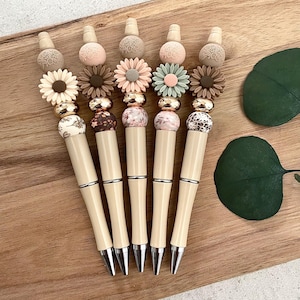 Pode incluir: Seis canetas bege com contas decorativas e pingentes em forma de flor. As contas t&ecirc;m v&aacute;rios padr&otilde;es, incluindo florais, estampa de leopardo e designs abstratos. Os pingentes em forma de flor s&atilde;o em tons de rosa, verde e marrom.