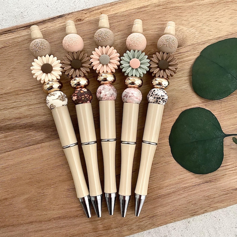 Pode incluir: Seis canetas bege com contas decorativas e pingentes em forma de flor. As contas t&ecirc;m v&aacute;rios padr&otilde;es, incluindo florais, estampa de leopardo e designs abstratos. Os pingentes em forma de flor s&atilde;o em tons de rosa, verde e marrom.