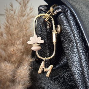 Peut inclure: Un mousqueton ovale doré fixé à un sac noir texturé. L'attache présente une breloque en forme de fleur beige avec une petite bande de strass et une breloque en forme de lettre M dorée suspendue au bas de l'anneau métallique ovale.