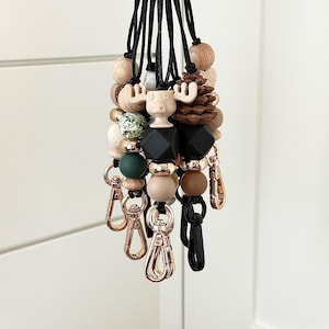 May include: A collection of wooden bead lanyards with various charms. The lanyards feature a moose, pinecone, and geometric black beads. The beads are in shades of brown, green, and cream, with gold-colored clasps.