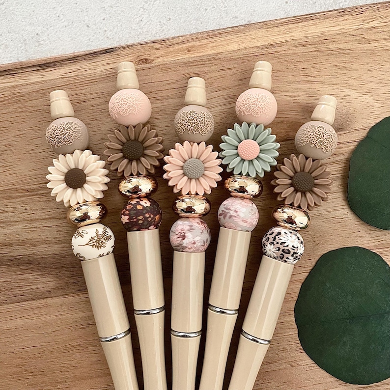 Pode incluir: Cinco canetas bege com contas decorativas. As contas s&atilde;o de v&aacute;rias cores e padr&otilde;es, incluindo desenhos florais, de estampa animal e geom&eacute;tricos. As canetas s&atilde;o coroadas com uma tampa bege.