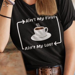 Peut inclure: Un t-shirt noir avec un graphique blanc qui dit "Ain't My First Ain't My Last" avec une tasse de café fumante entre le texte.