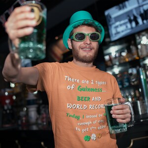 K&ouml;nnte beinhalten: Ein Mann mit einem gr&uuml;nen Hut und Sonnenbrille h&auml;lt zwei Gl&auml;ser mit gr&uuml;ner Fl&uuml;ssigkeit. Der Mann tr&auml;gt ein orangefarbenes T-Shirt mit dem Text "There are 2 types of Guinness... BEER AND WORLD RECORD. Today, I intend to drink enough of one to get the other."