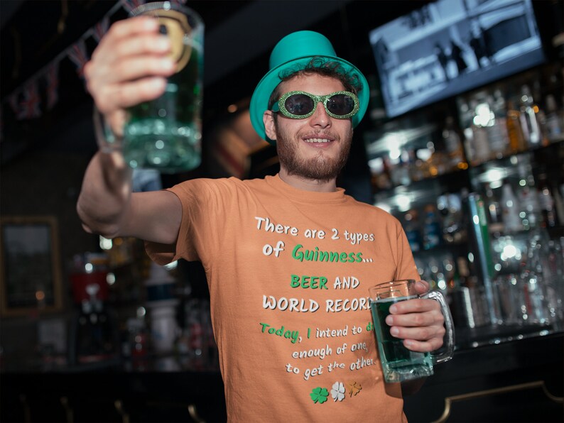 K&ouml;nnte beinhalten: Ein Mann mit einem gr&uuml;nen Hut und Sonnenbrille h&auml;lt zwei Gl&auml;ser mit gr&uuml;ner Fl&uuml;ssigkeit. Der Mann tr&auml;gt ein orangefarbenes T-Shirt mit dem Text "There are 2 types of Guinness... BEER AND WORLD RECORD. Today, I intend to drink enough of one to get the other."
