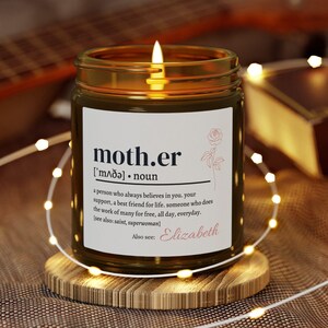 Puede incluir: Una vela encendida en un tarro de cristal con la palabra "mother" y su definición. La vela está sobre un posavasos de madera, rodeada de luces de cadena. El texto también incluye "Elizabeth".
