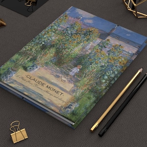 Può includere: Un taccuino con copertina rigida con un dipinto di Claude Monet di un sentiero di giardino con girasoli. La copertina include il testo "-CLAUDE MONET- I Must Have Flowers, Always and always." Matite dorate e nere poggiano su una superficie grigio scuro.