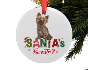 May include: A white, circular Christmas ornament featuring a Yorkshire Terrier. The ornament displays the text "SANTA'S Favorite" in red and green, with festive patterns and a Santa hat.