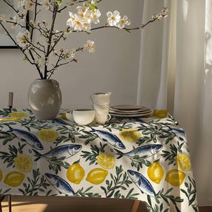 Puede incluir: Un mantel blanco con un patrón de limones y peces. El mantel está puesto sobre una mesa con un jarrón de flores blancas y algunos platos y tazas.