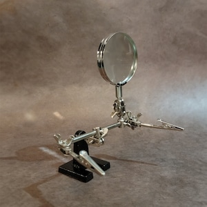 May include: A silver magnifying glass with a black base and adjustable arms. The magnifying glass is attached to a stand with two clamps.