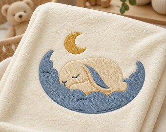 May include: A cream-colored plush blanket featuring an embroidered design of a light tan rabbit sleeping on a blue cloud. Above the rabbit, a small yellow crescent moon is stitched into the fabric. The blanket has a soft, fuzzy texture.