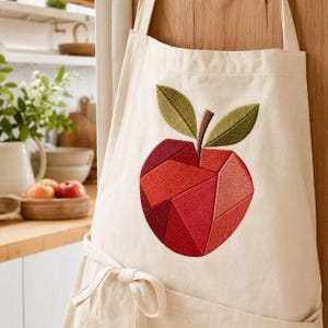 May include: A cream-colored apron featuring a large, embroidered apple design on the front. The apple is composed of various geometric shapes in shades of red and burgundy, topped with two olive green leaves and a brown stem, all stitched in thread.