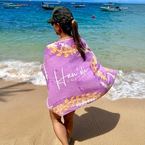 Puede incluir: Una persona en una playa arenosa con una toalla de playa morada con el texto "Hau'oli Anela" en blanco. La toalla tiene un borde floral amarillo y blanco. El fondo muestra un océano azul con botes y un cielo despejado.