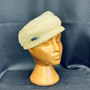 May include: A cream-colored straw hat with a tulle veil and a small black bug pin. The hat is on a wooden mannequin head.