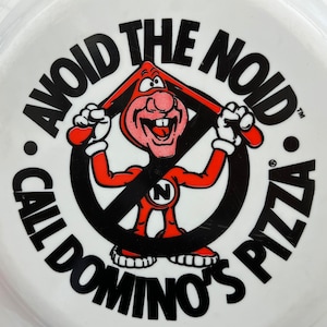 May include: A white circular sign with black text that reads "Avoid the Noid. Call Domino's Pizza."  A red cartoon character with a large nose and a black circle around him is in the center of the sign. The character is holding a large black circle with a white "N" in the center.