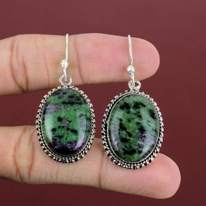May include: A pair of oval-shaped drop earrings featuring polished zoisite stones with green, black, and purple inclusions. Each stone is set in a silver-toned metal frame with a twisted rope-style border and attached to a simple silver hook wire.