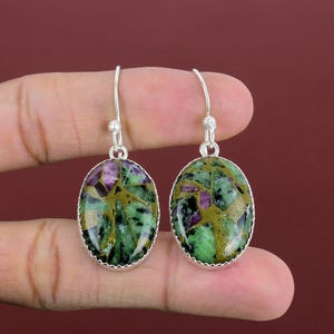 Peut inclure: Boucles d'oreilles ovales avec un sertissage argenté. Les boucles d'oreilles présentent une pierre verte et dorée avec des accents violets. Les boucles d'oreilles sont suspendues à un crochet simple.