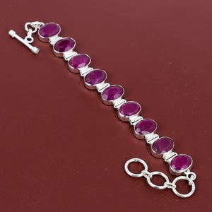 May include: A silver bracelet featuring nine oval-shaped, deep magenta-colored faceted stones set in individual metal frames. The piece includes a toggle clasp closure with a bar and a circular ring, connected by a series of small silver links.