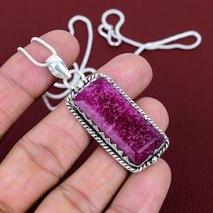 May include: A rectangular pendant featuring a vibrant magenta-coloured gemstone set in a silver-toned metal frame with a twisted rope-style border. The pendant hangs from a matching silver-toned chain, showcasing a faceted surface on the stone.