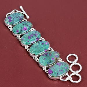 May include: A silver bracelet featuring six oval-shaped green and purple gemstones. The bracelet has a toggle clasp.