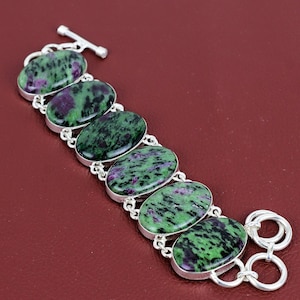 May include: A silver bracelet featuring oval-shaped gemstones with a green and purple pattern. The bracelet has a toggle clasp and is linked with silver connectors. The gemstones are approximately 1 inch in length.