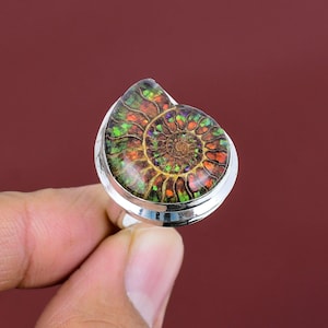 May include: A silver ring featuring a large, iridescent ammonite fossil. The fossil displays a spiral pattern with shades of green, orange, and brown. The ring is set against a deep red background.
