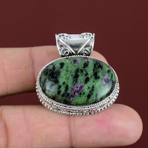 Puede incluir: Un colgante de forma ovalada con una piedra de zoisita pulida que presenta inclusiones verdes y negras vibrantes y parches de rubí magenta intenso. La piedra está engastada en un marco decorativo de plata esterlina con filigrana.