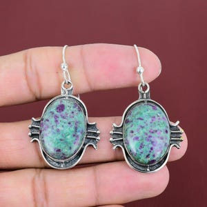 May include: A pair of silver earrings with oval-shaped green and purple gemstones. The earrings have a decorative silver frame and a hook closure. The gemstones have a speckled appearance.