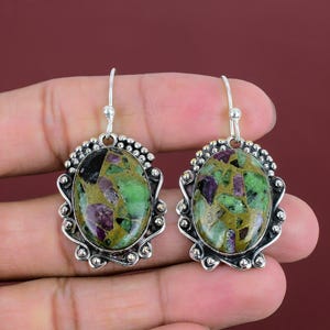 May include: A pair of silver earrings. Each earring features an oval gemstone with green, purple, and gold tones, set in a decorative silver frame. The earrings have a hook closure.