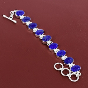 Puede incluir: Una pulsera de plata con piedras preciosas ovales de color azul oscuro. La pulsera tiene un cierre de palanca y está sobre un fondo granate. Las piedras están espaciadas uniformemente y conectadas por eslabones plateados.