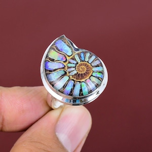 Op de afbeelding: Een zilveren ring met een groot, iriserend ammonietfossiel. Het fossiel heeft een spiraalpatroon met tinten blauw, groen en bruin. De ring wordt door een hand tegen een bordeauxrode achtergrond gehouden.