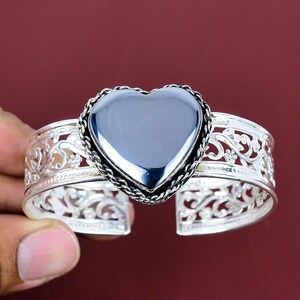 May include: A silver-toned cuff bracelet featuring an openwork filigree pattern and a large, smooth, dark gray heart-shaped stone centerpiece. The heart is framed by a twisted rope-style border, and the band has a wide, flexible open design.