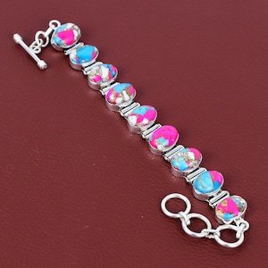 May include: A silver bracelet featuring oval-shaped stones in shades of pink, blue, and white. The stones are set in a silver metal band. The bracelet has a toggle clasp closure.