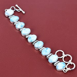 Könnte beinhalten: Ein silbernes Armband mit ovalen, hellblauen Edelsteinen. Das Armband hat einen Knebelverschluss und liegt auf rotem Hintergrund. Die Edelsteine sind durch silberne Glieder verbunden, was ein stilvolles Accessoire ergibt.