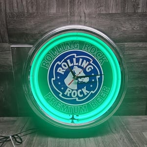 May include: A round, illuminated Rolling Rock Premium Beer clock. The clock face features a green and blue design with the Rolling Rock logo and text. The clock has a neon green border and a silver-colored frame.