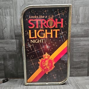 May include: Vintage Stroh Light Night sign with a gold frame. The sign features the words "Stroh Light Night" in red and gold lettering against a black background with a starburst design. A red and gold crest is at the bottom.