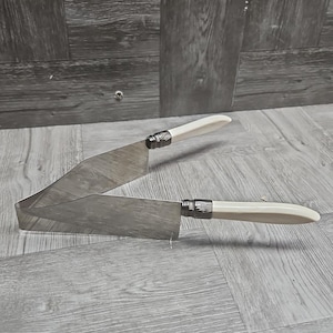 May include: Two stainless steel cake knives with ivory-colored handles and decorative silver-toned collars. The blades are angled, and the knives are displayed on a wooden surface.