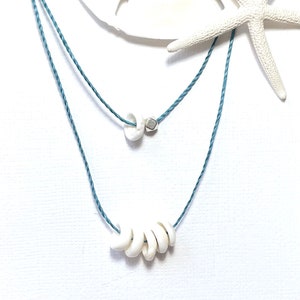 May include: A layered necklace featuring white shell beads and a small silver-colored bead strung on a light blue cord. The necklace is displayed with a white starfish and a sand dollar.