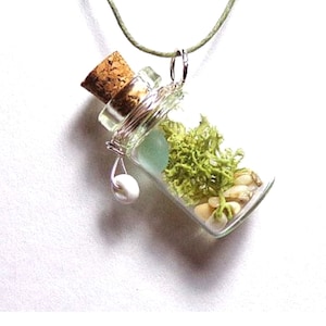 May include: A glass bottle necklace with a cork top, filled with green moss, white seashells, and a light blue sea glass piece. The bottle is suspended from a green cord.